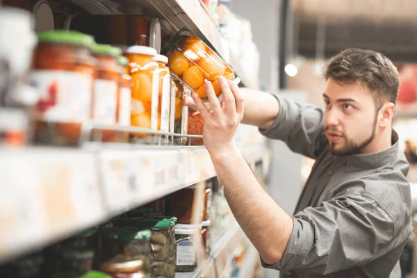Adult man takes a can of vegetables with a shelf from a grocery store. Choosing and buying canned tomatoes in a supermarket. Man buys products in a supermarket, takes from a shelf. — Stock Photo, Image