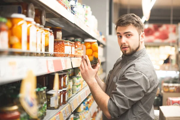 Ritratto di un bellissimo uomo con la barba in piedi nella navata del supermercato, un reparto di cibo in scatola, con in mano un barattolo di verdure in scatola e guardando nella macchina fotografica. — Foto Stock