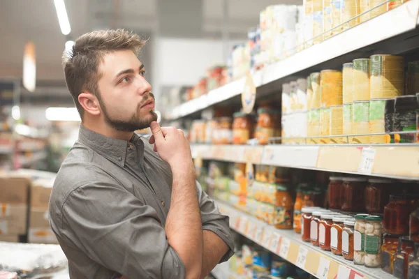 Koper man kijkt naar de supermarkt plank en denkt welk product te nemen. Koper selecteert ingeblikt voedsel in de winkel. Winkelen in een supermarkt. — Stockfoto