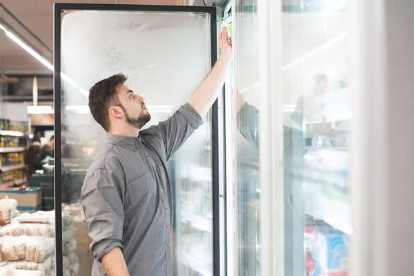 Pembeli memecahkan pintu kulkas di supermarket dan mengambil produk rak beku. Seorang pria memilih makanan beku di lemari es di toko kelontong. Belanja dalam konsep supermarket . — Stok Foto