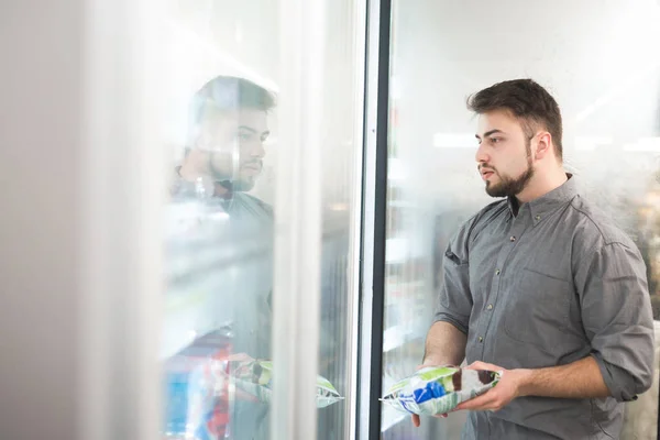 Portrait d'un homme debout près du réfrigérateur dans un supermarché avec un paquet d'aliments surgelés à la main et regardant l'étagère dans le réfrigérateur.L'acheteur sélectionne des aliments surgelés à l'épicerie — Photo