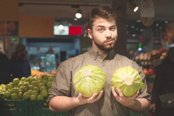 Retrato de un hombre parado en el departamento de verduras de un supermercado con dos repollos en las manos, mirando a la cámara. ? abbage comprador en un supermercado . — Foto de Stock