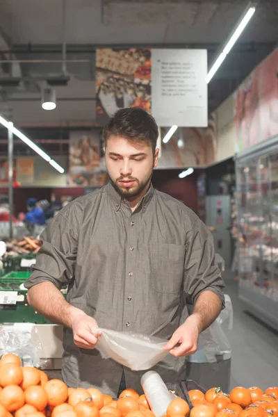 Male customer holding fresh orange and choosing citruses. Bearded man standing in supermarket in fruit section and do shopping. Man holding red basket full of products. — Stock Photo, Image