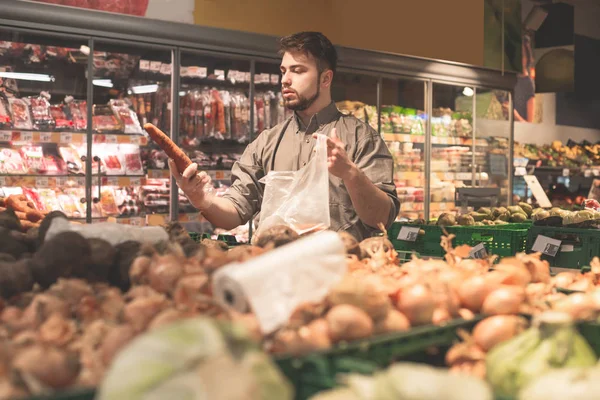 Portrait of a man chooses vegetables in a supermarket. Holds a carrot his hand and puts it in a package. Bearded buyer a man buys vegetables in a supermarket. — Stock Photo, Image