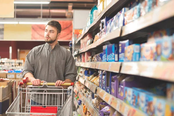 Portretul unui om cu un cărucior stă într-un supermarket din culoar lângă rafturi cu produsele și se uită la cameră. Portretul unui cumpărător într-un supermarket . — Fotografie, imagine de stoc