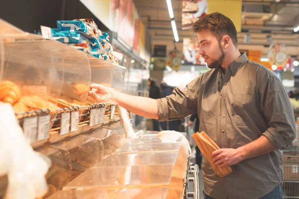 Man buys food in the bread department of the supermarket.Man a shirt holds a paper bag in his hands, stands in the bread department of the supermarket and selects buns. Buyer buys bakery products