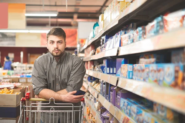 Porträt eines glücklichen Mannes, der in einem Einkaufswagen auf dem Gang eines Supermarktes steht, in die Kamera blickt und lächelt. macht Einkäufe Supermarkt — Stockfoto