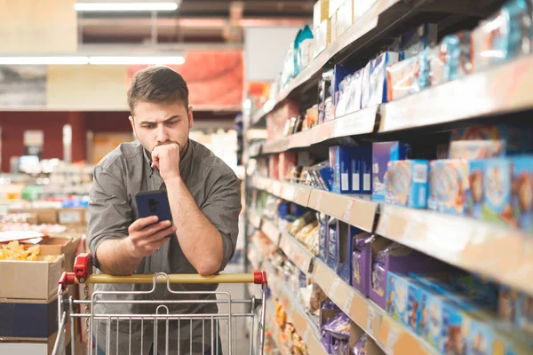 El hombre serio está parado en un carro el pasillo de un supermercado, mirando la pantalla de un smartphone.Buyer un supermercado utiliza un teléfono inteligente. Hombre en un teléfono inteligente en las manos de una tienda de comestibles — Foto de Stock