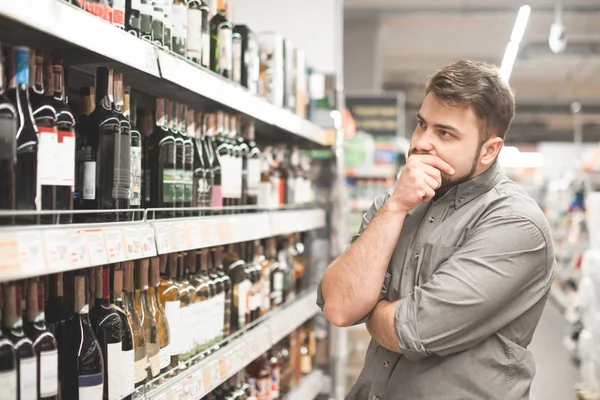 Elegante hombre afroamericano casual en jeans chaqueta y boina negra celebración de la cesta y mirando en la botella de vino, compras en el supermercado . — Foto de Stock
