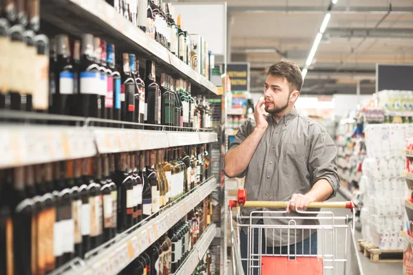 Mann geht auf ein Regal und wählt Wein. Der Käufer läuft mit einem Einkaufswagen durch den Supermarkt, betrachtet das Weinregal. bärtiger Mann wählt und kauft Wein. — Stockfoto