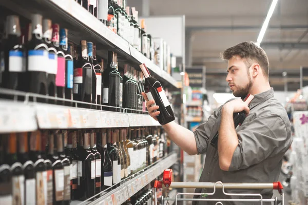 Esto debería estar bien. Vista lateral de un joven guapo sosteniendo una botella de vino y mirándola mientras está de pie en una tienda de vinos — Foto de Stock