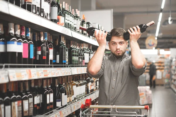Bir adamın portresi bir süpermarkette şarap satın alır, iki şişe tutar ve ne seçmez, kameraya bakar. Bir süpermarkette bir alıcının portresi kafasından şarap şişeleri tutuyor — Stok fotoğraf