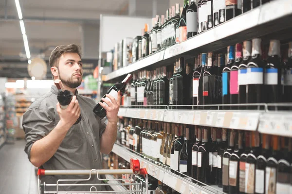 Koper met een kar staat in de alcohol-afdeling van de supermarkt in de buurt van de planken met wijn, twee flessen wijn in zijn handen houdt, kiest wat te kopen. Man kiest en koopt wijn in een supermarkt — Stockfoto
