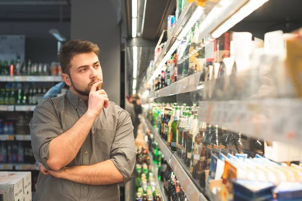 Business man is een koper staande in een supermarkt en het kiezen van een drankje. Attente man met een baard kiest bier in een supermarkt, richt zich op bierflessen en denkt. Koper koopt een biertje in een supermarkt — Stockfoto