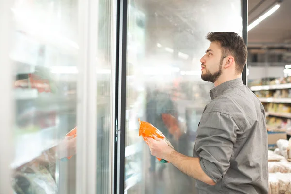 Man met een baard staat bij de koelkast in een supermarkt met een bevroren product in zijn handen en kijkt naar de plank. Koper selecteert diepvriesproducten in de supermarkt. — Stockfoto