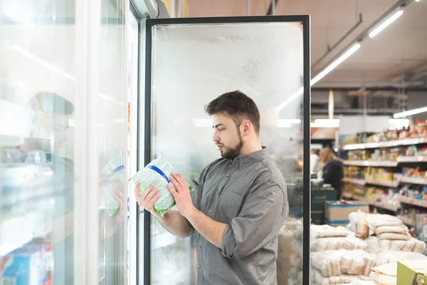 O comprador quebra a porta do frigorífico no supermercado, segura a embalagem com as mãos e olha para o rótulo. O homem escolhe comida congelada no frigorífico do supermercado. Escolha de alimentos congelados no supermercado . — Fotografia de Stock