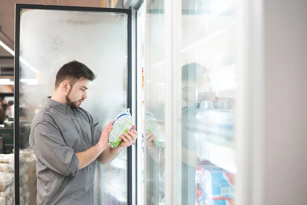 El comprador de pie en el supermercado cerca del refrigerador sostiene el paquete en su mano y lee la etiqueta. El hombre comprueba la fecha de producción de alimentos congelados en el supermercado . —  Fotos de Stock