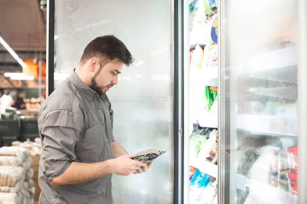 Homem barbudo com comida congelada suas mãos estão na geladeira no departamento de alimentos congelados. Comprador seleciona e compra alimentos congelados no supermercado. Compras em um supermercado . — Fotografia de Stock