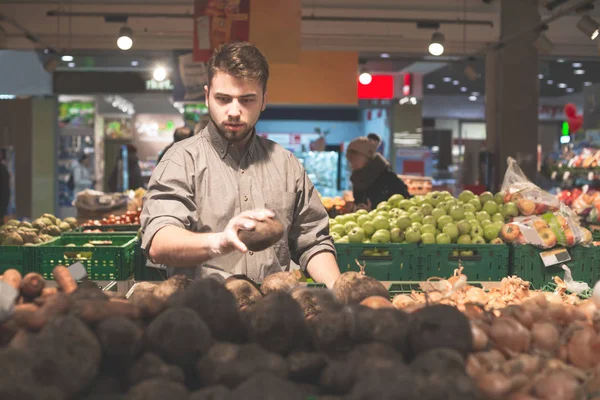 El hombre con manoplas selecciona verduras en un supermercado. Hombre adulto cavando ingredientes para platos en una tienda de comestibles, un departamento de verduras. Comprador compra verduras . — Foto de Stock