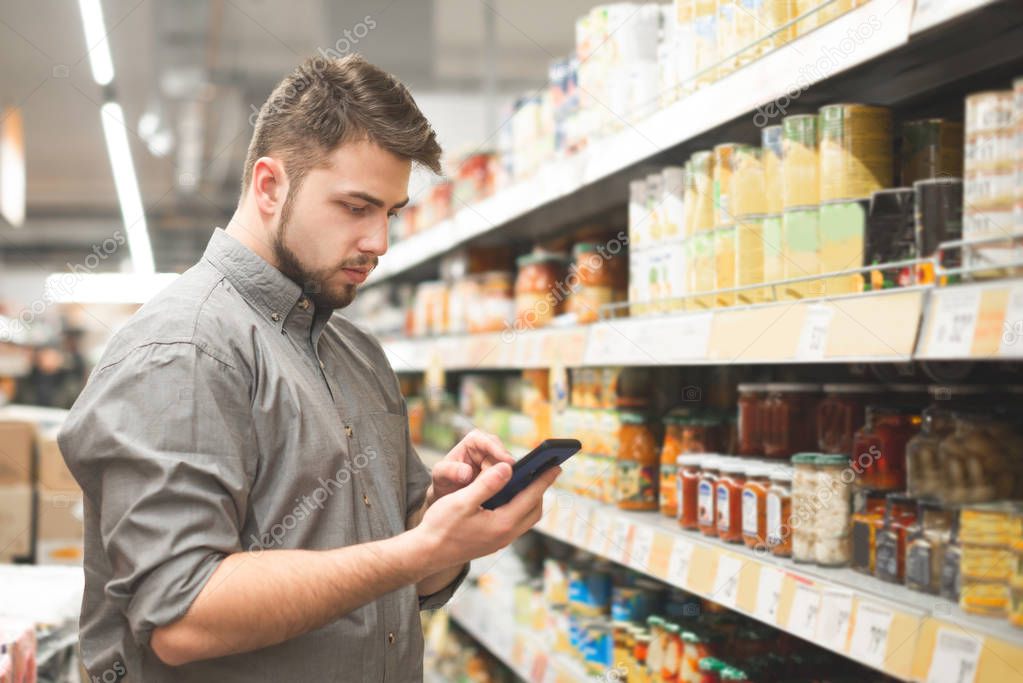 Man stands in the aisle of the supermarket at shelves with canned vegetables and uses a smartphone. Buyer man looks at the shopping list on the smartphone and selects the products in the supermarket