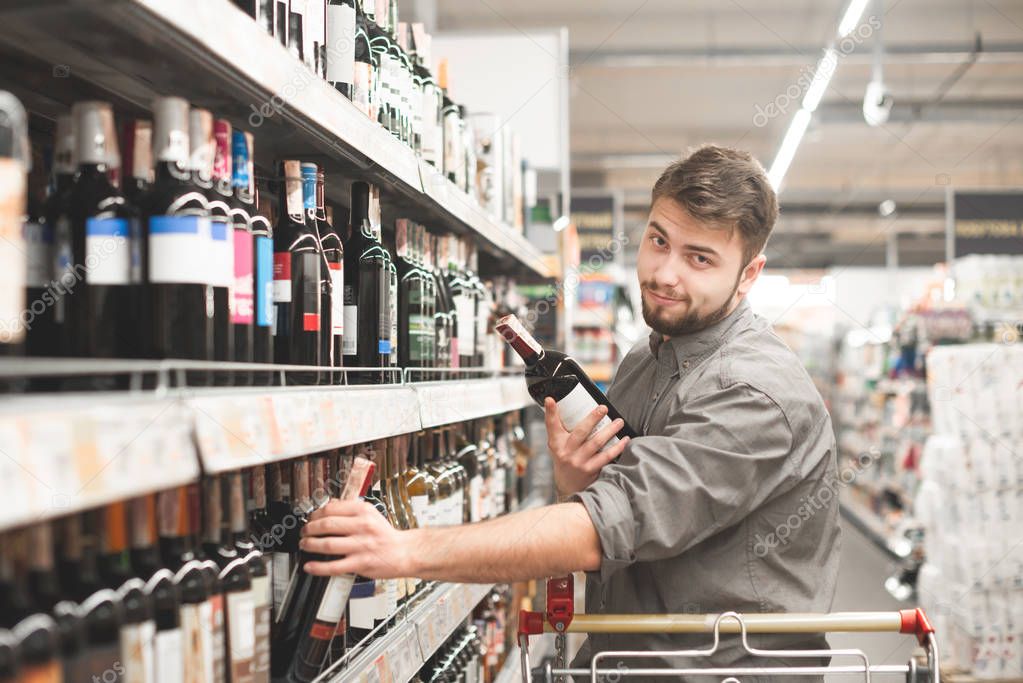 Cheerful man buys wine at a grocery store. Funny man with bottles of wine in the alcohol department of the supermarket. Positive buyer takes the wine out of the shelves and looks at the camera.