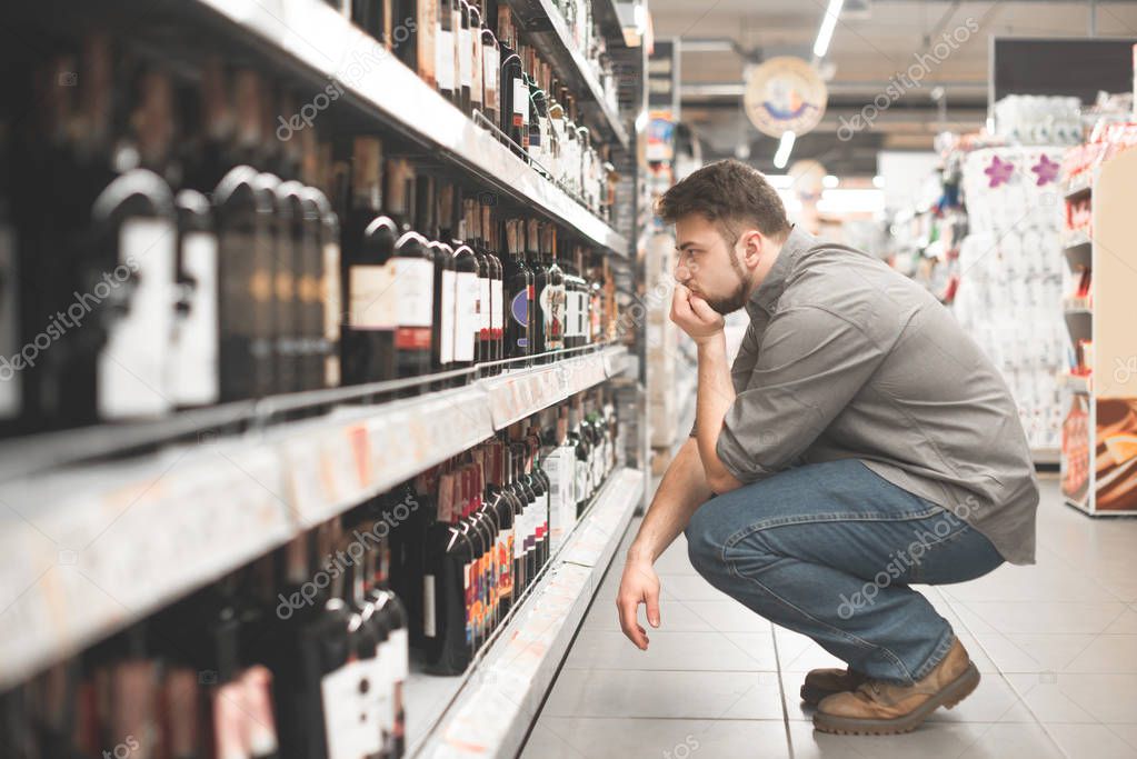 Man sitting on the aisle in the supermarket and look at the shelves of wine, Buyer chooses wine in a supermarket. Choosing and buying wine in the supermarket.