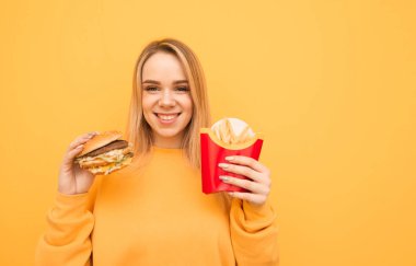 Elinde Burger ve patates kızartması ile rahat bir turuncu giyim gülümseyen çekici kız sarı bir arka plan üzerinde duruyor, kamera bakıyor ve gülümsüyor. Gülümseyen kız ve Fast Food.