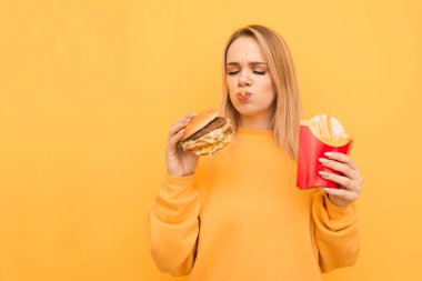 Ağzına patates kızartması ile aç komik kız sarı bir arka planda elinde Burger bakıyor. Turuncu kıyafetler neşeli kız lezzetli Fast Food, izole yiyor. Zararlı gıda.