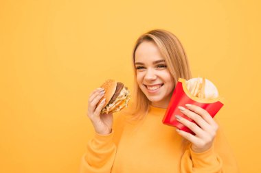 Sarı bir arka planda elinde Burger ve patates kızartması ile yakın portre gülümseyen sarışınlar, görünümlü kamera ve gülümseyen, turuncu giysiler giyiyor. Hızlı gıda ile mutlu kız, sarı bir izole