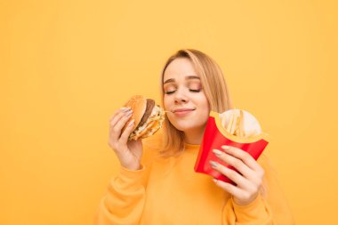 Bir kızın yakın çekim portresi, gözleri kapalı, kızarmış patates ve sarı arka planda bir burger yeme ile hızlı yemek sever. Sarı bir arka planda izole çekici sarışın kalori Gıda. Kopya-boşluk