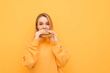 Turuncu kıyafetler komik kız sarı bir duvar arka planda bir burger yiyor, ısırır ve kameraya bakar. Aç kız öğrencisi kötü yemek yiyor, ısırıyor. Izole.