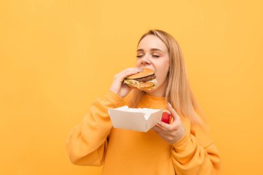 Bir kızın gözleri sarı bir arka planda kapalı bir burger ısırma portre closeup, turuncu giyim giyiyor. Aç kız, zararlı bir yiyecek yiyor ambalaj ve Burger tutar.
