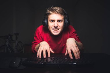 Funny young man typing text on the computer with two hands and focusing on the computer screen, sitting at the table and playing video games. Focused teenager is playing video games on a computer clipart