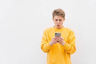 Casual giyim hayran genç adam beyaz bir duvarın arka planda duruyor ve bir akıllı telefonun ekrana bakar. Şok adam bir smartphone kullanır. Açık duvarın arka planında sokak portresi