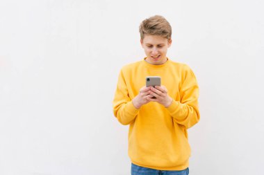 Sarı bir sweatshirt bir adam yakın çekim portre, beyaz bir duvara ayakta, bir akıllı telefon sevinçli zevk. Mutlu genç Casual giyim akıllı telefonda bir mesaj yazar ve gülümsüyor