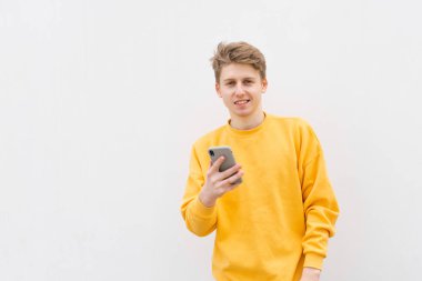 Beyaz bir duvara karşı elinde bir smartphone ile pozitif genç adam, sarı bir sweatshirt giyiyor, kameraya bakarak ve gülümsüyor. Öğrenci elinde akıllı telefon tutan, sokak portre.