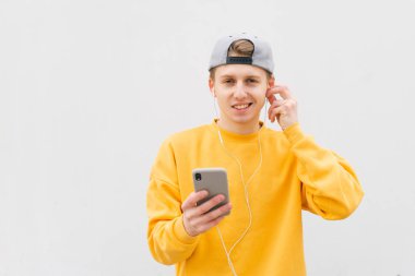 Beyaz bir duvarın arka planına karşı elinde bir smartphone ile ayakta neşeli adam ve kulaklık müzik dinlerken, sarı bir sweatshirt giyiyor, kamera bakarak ve gülümseyen
