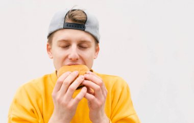 Beyaz bir duvarın arka planına karşı gözleri kapalı bir sandviç yiyen genç bir adamın yakın çekim portresi. Aç öğrenci beyaz bir arka planda bir burger yiyor. Boşaltmak
