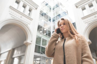 Genç iş kadını, modern kentsel mimarinin arka planında telefonda arıyor, uzağa bakıyor. Şehir arka plan, modern mimari ve iş kızı telefonda konuşan bir ceket kız