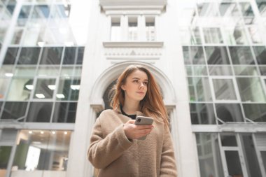 Modern bir kentsel binanın arka planına karşı elinde bir smartphone ile güzel kız ayakta, uzak bakıyor, şık bayan bir ceket giyiyor, kentsel arka planda bir akıllı telefon kullanarak.