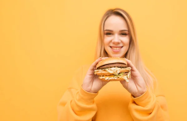 Happy girl with a burger in her hands on an orange background, looking into the camera and smiling, wearing yellow clothes. Focus on appetizing burgers in the hands of a smiling girl.Closeup photo