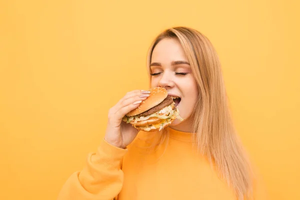 Bir kızın gözleri sarı bir arka planda kapalı bir burger ısırma portre closeup, turuncu giyim giyiyor. Aç kız bir zararlı gıda yiyor, büyük bir lezzetli burger elinde tutar. — Stok fotoğraf