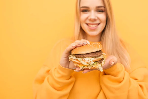 자른 사진, 행복한 소녀는 그녀의 손에 좋은 맛있는 햄버거를 들고, 오렌지 배경에 고립 된 카메라를 보고 미소를 지었습니다. 그녀의 손에 햄버거와 미소 소녀, 손에 초점. — 스톡 사진
