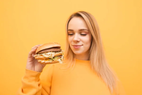 그의 손에 햄버거와 매력적인 금발은 패스트 푸드를 응시, 노란색 배경에 서있다. 배고픈 소녀는 노란색 배경에 그녀의 손에 햄버거를 들고있다. 햄버거에 초점. — 스톡 사진