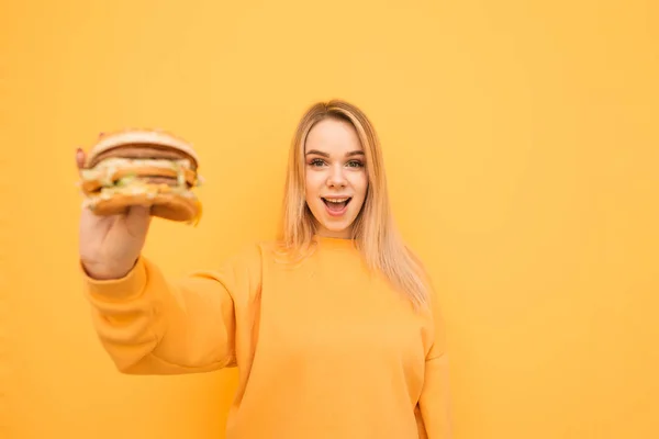 Muchacha atractiva con una cara sorprendida se encuentra sobre un fondo amarillo en una ropa informal naranja, sosteniendo una deliciosa hamburguesa sabrosa en la mano.Chica alegre con comida rápida en las manos se aísla en un amarillo — Foto de Stock