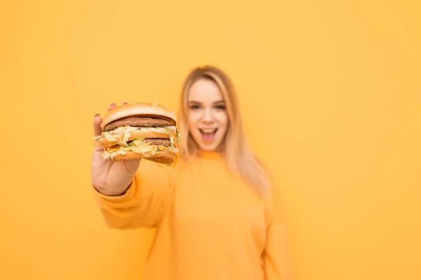 Menina feliz em uma roupa casual laranja detém um grande hambúrguer saboroso em suas mãos e mostra-lhe em uma câmera em um fundo amarelo. Concentre-se no hambúrguer nas mãos de uma rapariga expressiva. Espaço de cópia — Fotografia de Stock