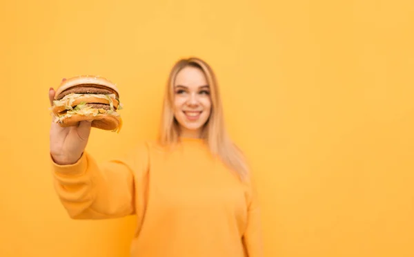 Foto de um hambúrguer apetitoso em sua mão em uma menina sorridente feliz fica em um fundo amarelo, vestindo roupas casuais laranja. Comida rápida nas mãos de uma rapariga. Conceito de alimento nocivo . — Fotografia de Stock