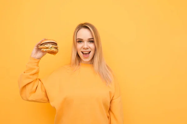 오렌지 색 캐주얼 의상을 입고 노란색 배경에 햄버거를 들고 포즈를 취하고 카메라를 바라보며 미소를 짓고 있는 행복한 소녀. 미소 짓는 소녀는 그녀의 손에 좋은 맛있는 햄버거의 손에 들고 — 스톡 사진