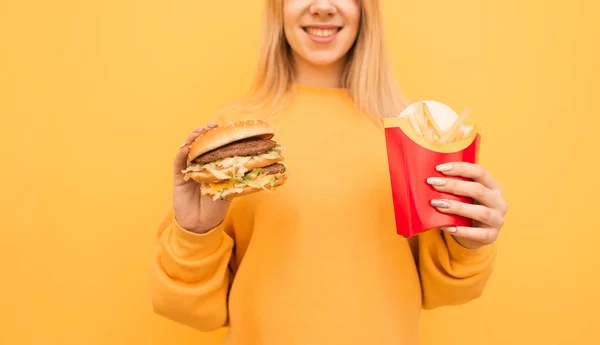 Happy young girl in orange sweatshirt with burger and fries in hands, smiling, isolated on yellow background, cropped close up photo. Copyspace — ストック写真