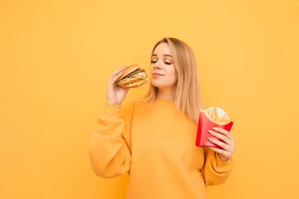 매력적인 금발 소녀는 그녀의 손에 유해한 음식과 노란색 벽의 배경에 서서 갈 것입니다. 소녀는 그의 손에 감자 튀김을 들고, 신선한 햄버거의 냄새를 즐긴다. 격리 — 스톡 사진